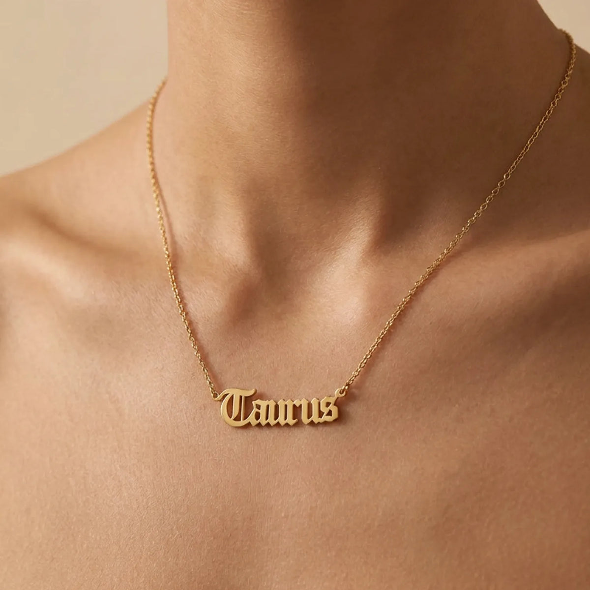 A gold nameplate necklace with a gothic spelling 'Taurus' worn on a model's neck, shown in a warm, close-up shot against a neutral beige background.