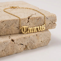 A gold nameplate necklace spelling 'Taurus' in a gothic font, resting on stacked beige stone blocks against a white background.