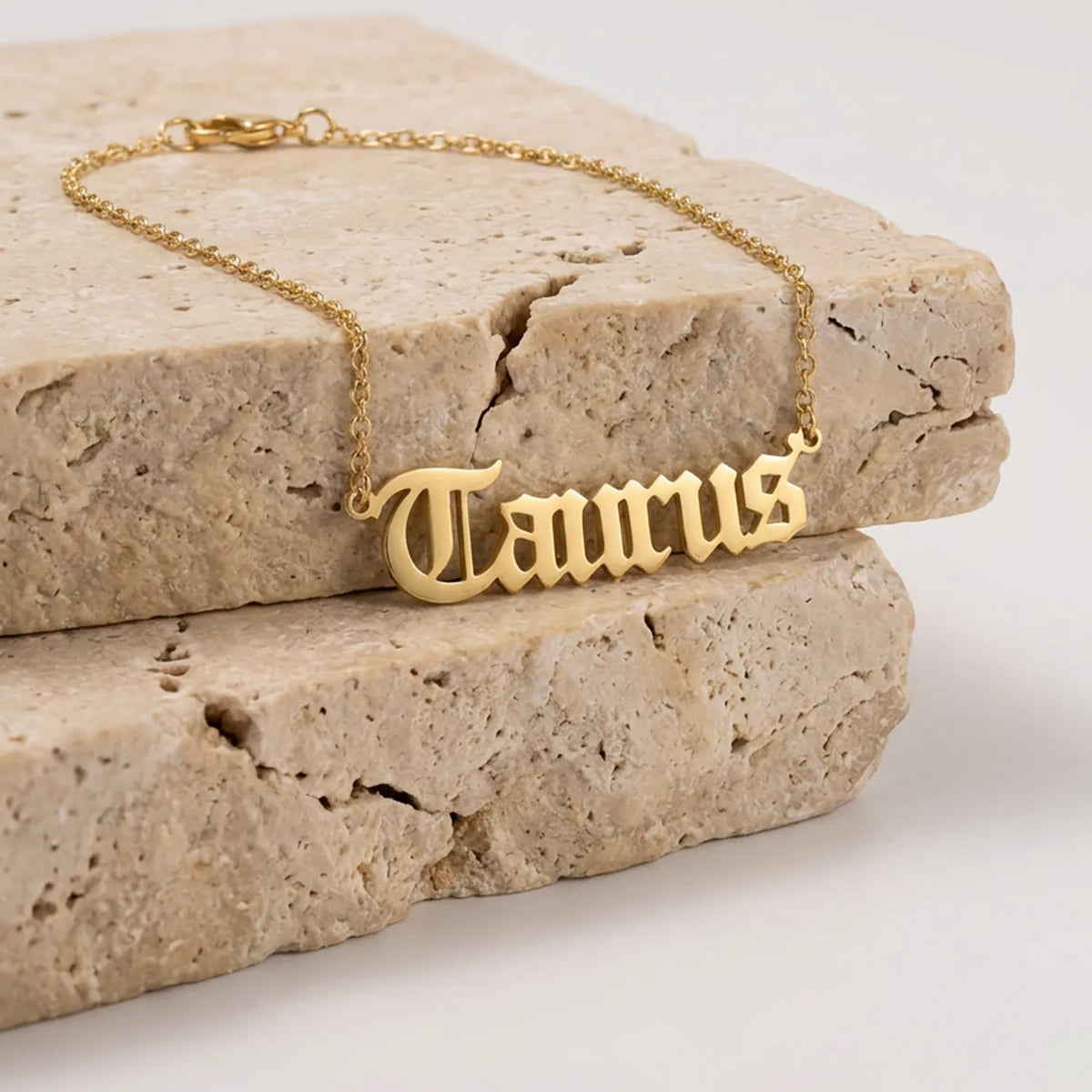 A gold nameplate necklace spelling 'Taurus' in a gothic font, resting on stacked beige stone blocks against a white background.