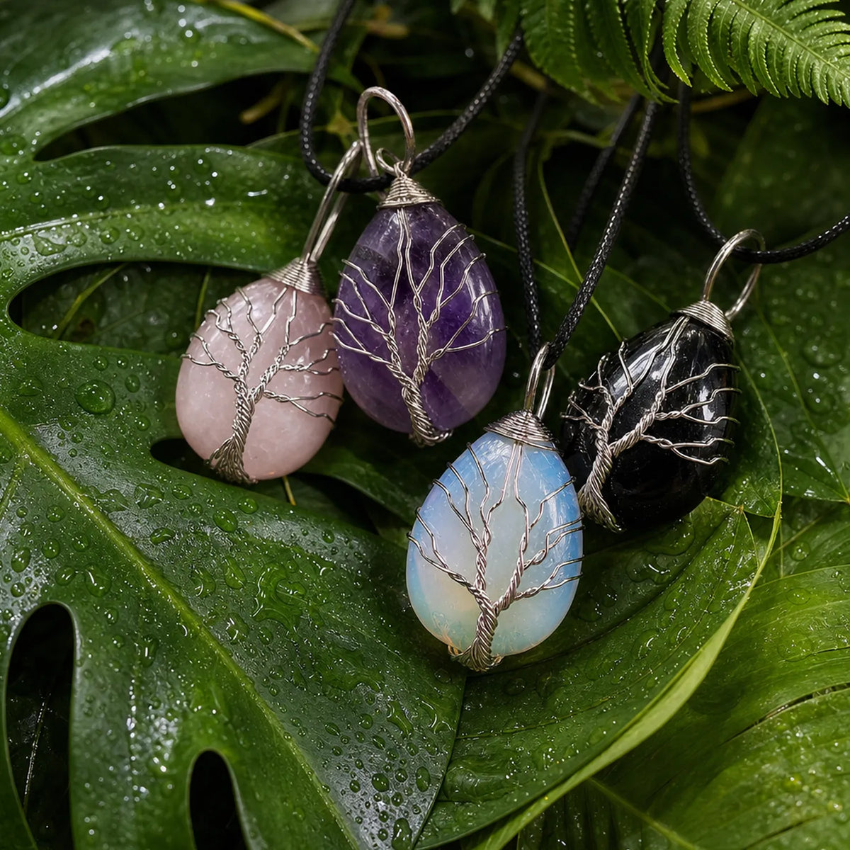 Four teardrop-shaped crystal pendants with tree-of-life wire designs hang on black cords against a backdrop of wet green leaves. The pendants are made from pink, purple, light blue, and black stones.