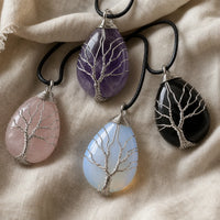Four teardrop-shaped crystal pendants with silver tree-of-life wirework, shown on black cords against a beige linen fabric background. The stones are rose quartz, amethyst, opalite and 