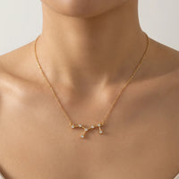 A delicate gold necklace with a Virgo constellation pendant featuring clear gemstones, shown worn on a model's neck against a soft beige background.