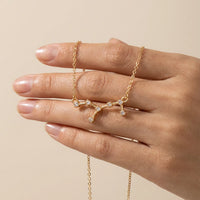 A gold chain necklace with a virgo constellation pendant set with small clear stones, shown worn on a hand against a soft beige background.ion