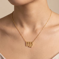 A close-up of a model wearing a delicate gold chain necklace with a zodiac sign pendant, specifically the Libra symbol, against a soft beige background.