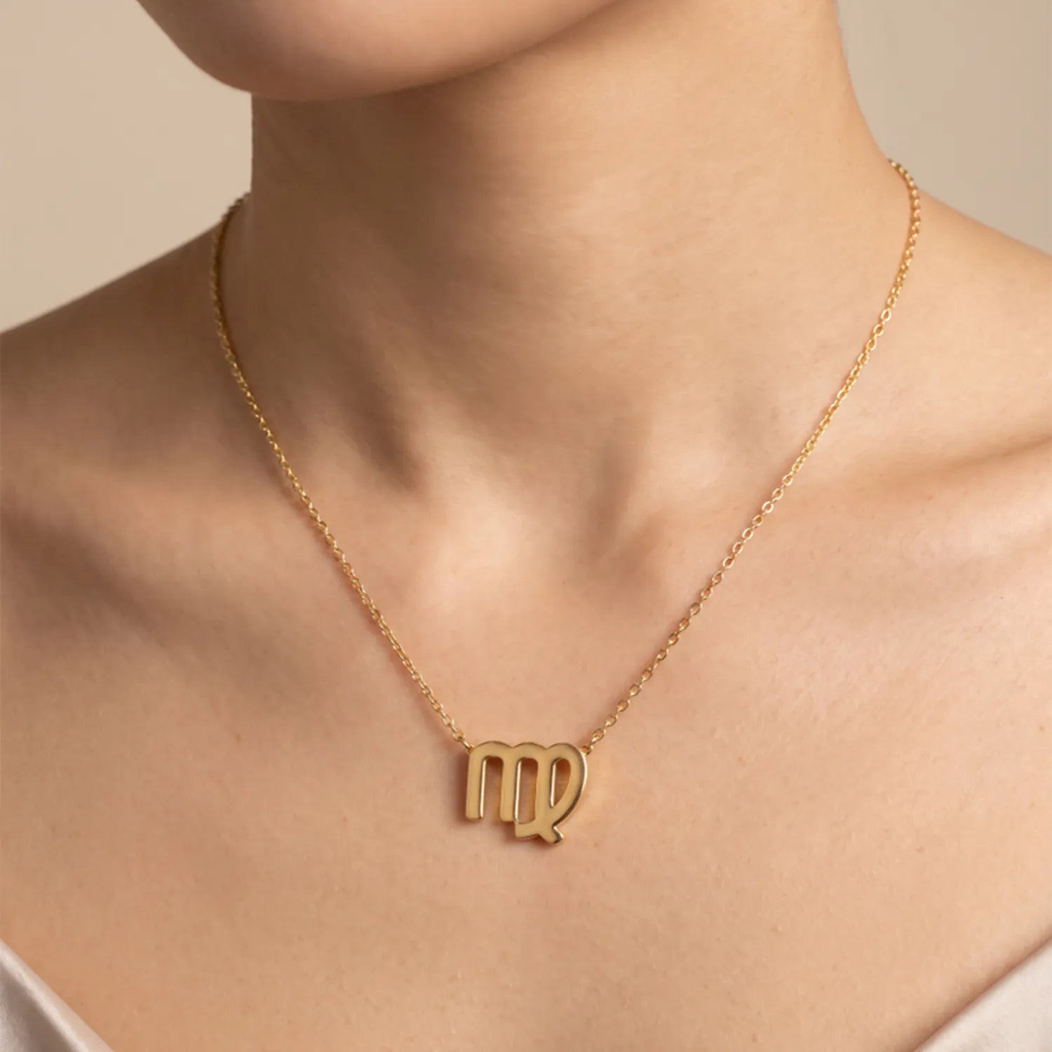 A close-up of a model wearing a delicate gold chain necklace with a zodiac sign pendant, specifically the Libra symbol, against a soft beige background.