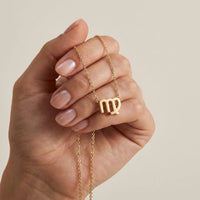 A close-up of a hand holding a delicate gold chain necklace with a gold Virgo pendant against a soft beige background.