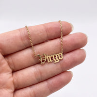 A gold nameplate necklace spelling 'Virgo' in a gothic font, held on a person's fingers against a clean white background.
