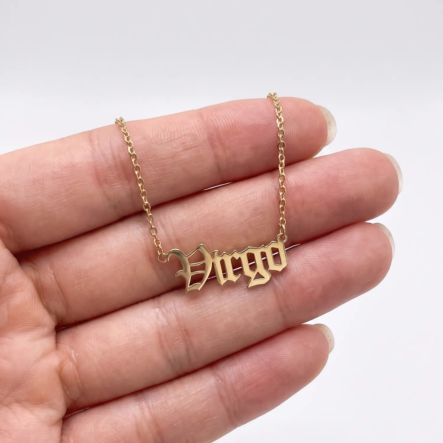 A gold nameplate necklace spelling 'Virgo' in a gothic font, held on a person's fingers against a clean white background.