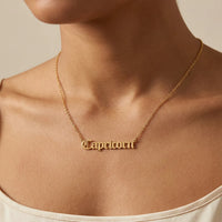 A gold nameplate necklace spelling 'Capricorn' in a gothic script worn on a model's neck, shown in a close-up shot against a warm beige background.