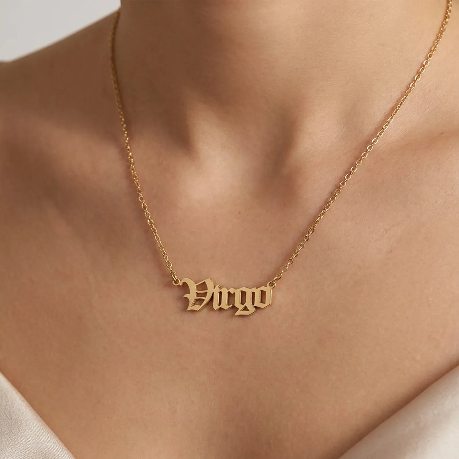 A gold nameplate necklace spelling Virgo in a gothic font, worn on a model's neck over a white top.